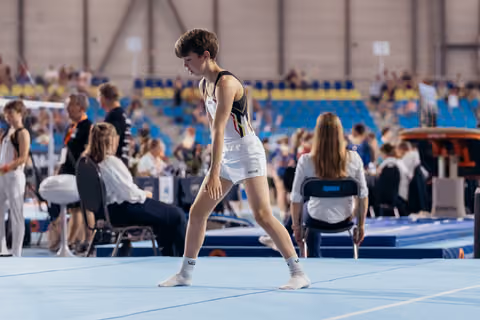 Young gymnast in Belgian colours stands composed on the blue floor, gathering focus before or after a routine.