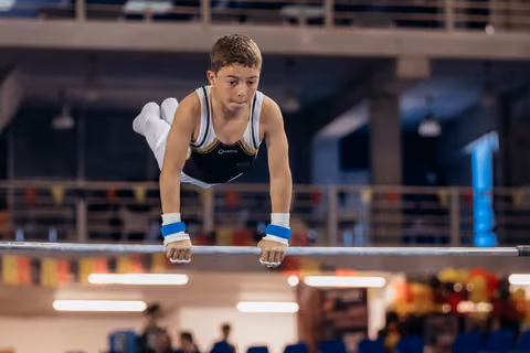 Focused teenage gymnast performs a support hold on parallel bars, wearing blue wristbands, with intense concentration.