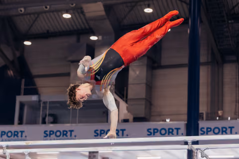 Young male gymnast in red and black leotard performs inverted release on parallel bars at indoor sports hall.
