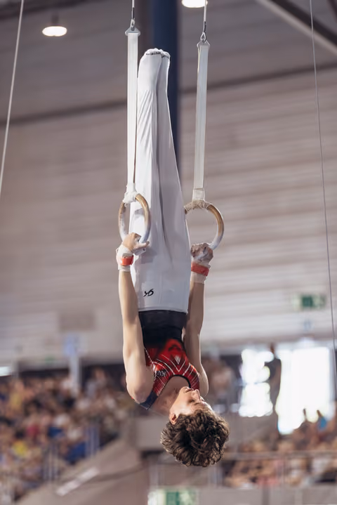 Young male gymnast hangs inverted on still rings, expression calm and focused, performing in a packed indoor arena.