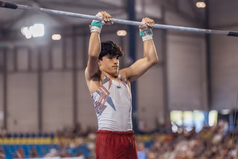 Young male gymnast grips the high bar with intense focus, wearing a white leotard and wrist guards in an indoor arena.