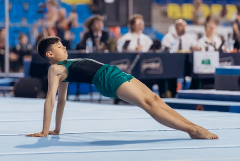 Young male gymnast performs a graceful floor routine, holding a split-leg pose with focused expression at a gymnastics meet.