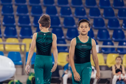 Two young male gymnasts in green Quatro uniforms walk together in an arena, one facing forward with a focused expression.