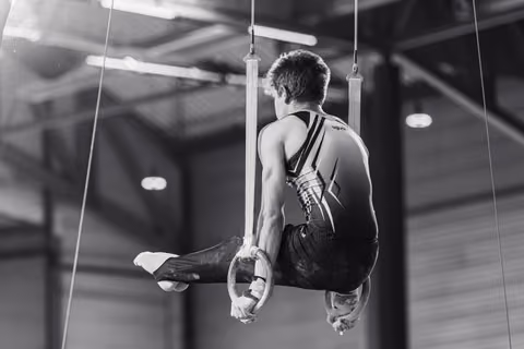 Male gymnast performs on still rings in a tuck position, showing focused strength and controlled technique in an indoor gymnasium.