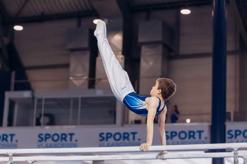 Young male gymnast performs a strong horizontal hold on parallel bars, legs raised high, at a Sport Vlaanderen event.