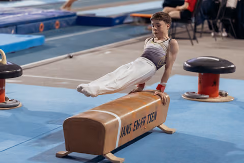 Young male gymnast performs a scissor hold on the pommel horse, concentrated expression, in a blue gymnasium.