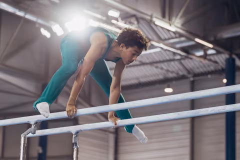 Young male gymnast in teal leotard leans forward on parallel bars with intense concentration in an indoor gymnasium.