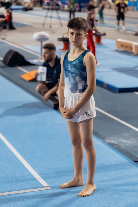 Young male gymnast stands barefoot on blue floor, hands clasped, composed expression, awaiting his turn at indoor gymnastics meet.