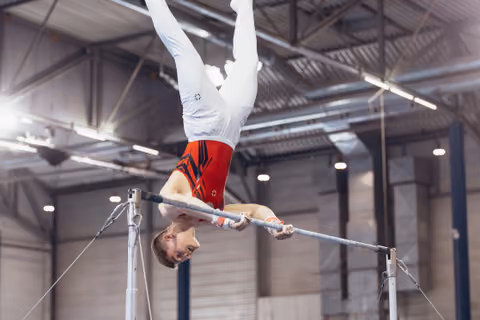 Male gymnast holds an inverted position on the high bar, body fully extended upward, focused expression, in a bright indoor gymnasium.