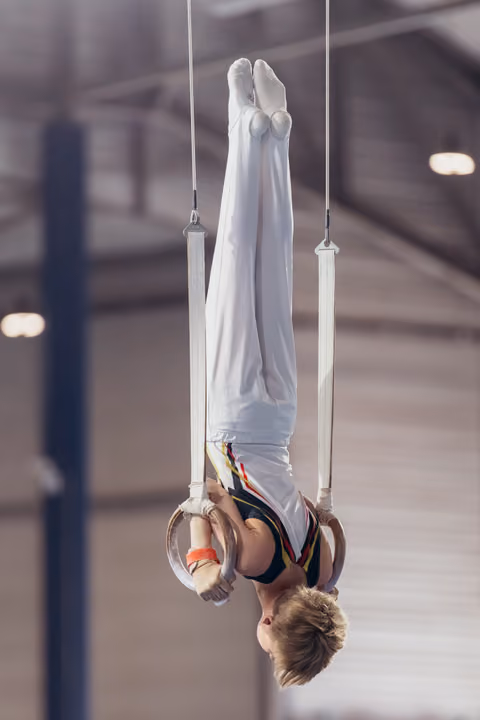 Young male gymnast performs an inverted hang on still rings, showing strength and control in a sports hall.