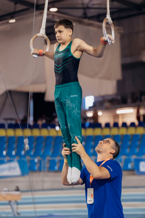Focused young gymnast holds still rings while coach steadies his legs in a blue-seated gymnastics arena.
