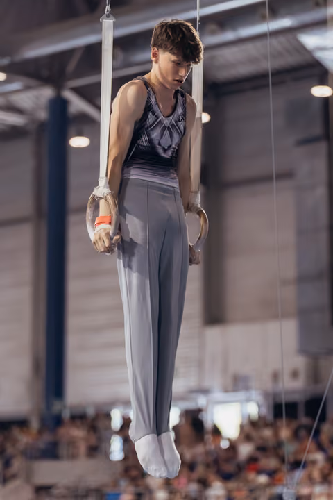 Young male gymnast hangs on still rings with intense focus, wearing gray competition pants in a packed arena.