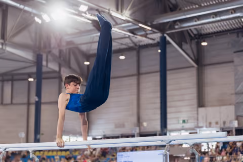 Young male gymnast executes a powerful pike hold on parallel bars, legs raised high, intense focus in an indoor arena.