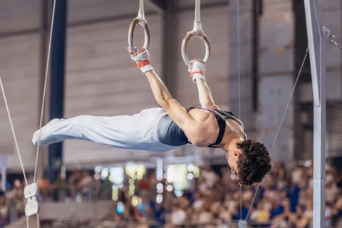 Male gymnast executes a powerful inverted hang on still rings, body perfectly horizontal, showing intense concentration before a crowd.