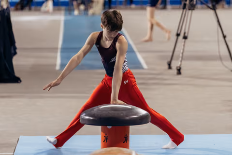 Young male gymnast performs a straddle vault, leaning forward with intense focus during a gymnastics competition.