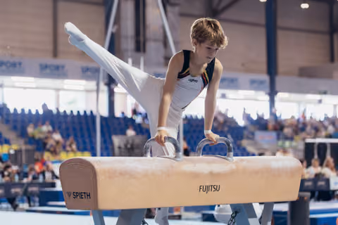Young male gymnast performs on pommel horse, leg raised high, focused expression, indoor arena with spectators.