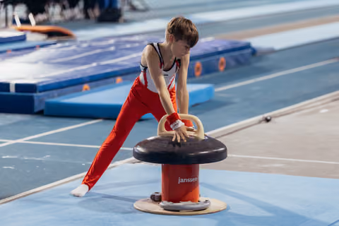 Young male gymnast leans intently over a mushroom apparatus, gripping the handles with focused concentration during competition.