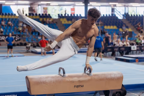 Young male gymnast performs scissors on a Fujitsu pommel horse, legs split wide, intense concentration on his face.