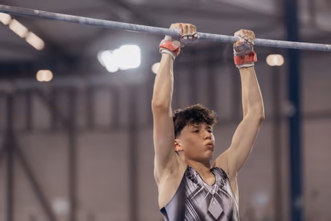 Young male gymnast hanging from the high bar with intense focus, wearing red grips and a geometric leotard indoors.