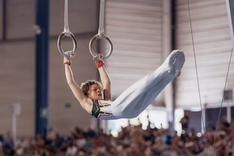 Young male gymnast performs on still rings, legs raised in an L-shape, focused expression, competing in a packed indoor gymnasium.