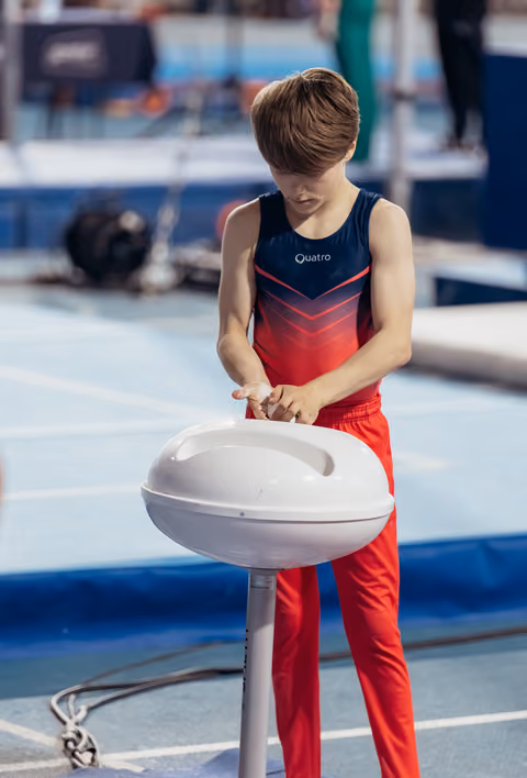 Young male gymnast in red and navy leotard applies chalk to hands at a chalk bowl, focused before his routine.