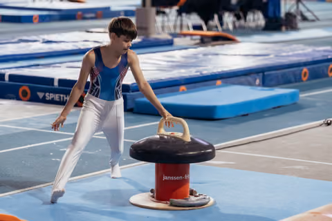 Young male gymnast in blue leotard leans forward intently, gripping a vault pommel horse during training at a gymnastics meet.