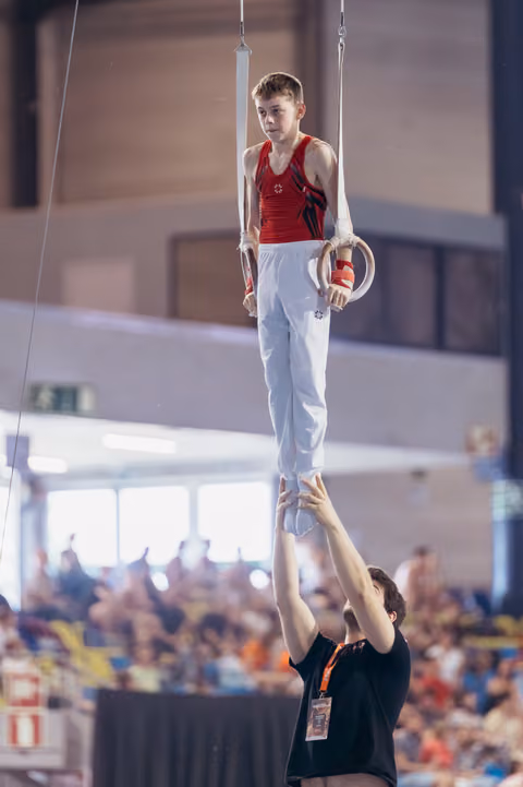 Young male gymnast holds still rings with intense focus while coach below steadies his feet in a packed arena.