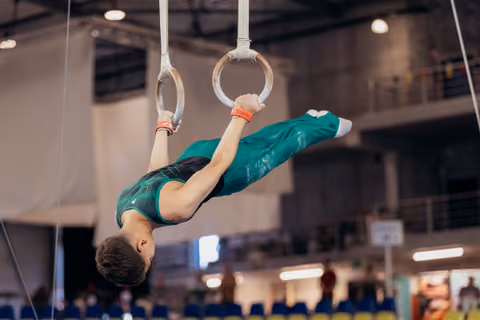 Young male gymnast holds an inverted position on still rings, body horizontal, wearing a teal leotard in a competition hall.