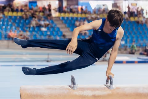 Young male gymnast performs pommel horse routine, legs split mid-air, concentrated expression in arena setting.
