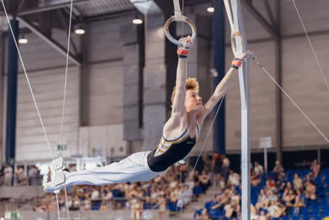 Young male gymnast performs a powerful horizontal hold on rings, body perfectly parallel, focused expression, in a packed arena.