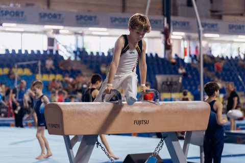 Young gymnast in Agiva kit concentrates intently on the pommel horse during an indoor gymnastics meet.