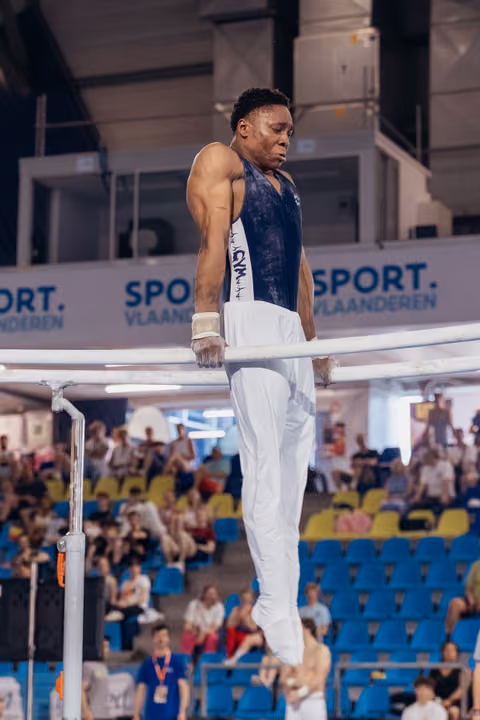 Male gymnast grips parallel bars with intense focus, muscular arms engaged, competing before a packed arena crowd.