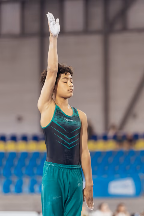 Young male gymnast stands with eyes closed, one arm raised in salute, wearing teal and black leotard in arena.