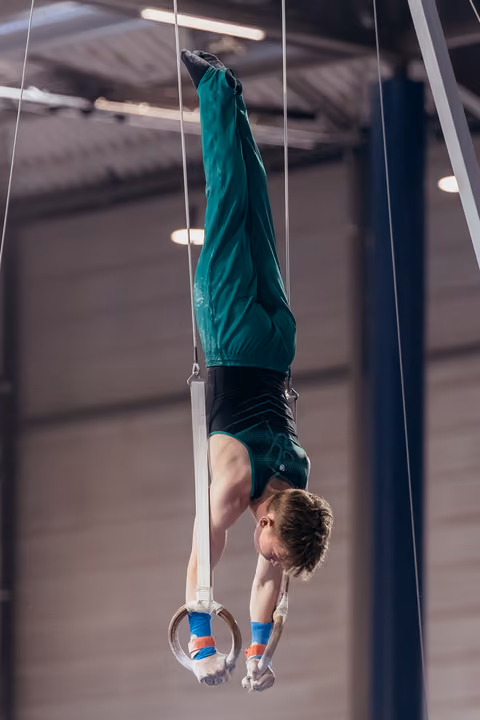 Young male gymnast performs an inverted hang on still rings, body fully extended upside down in a sports hall.