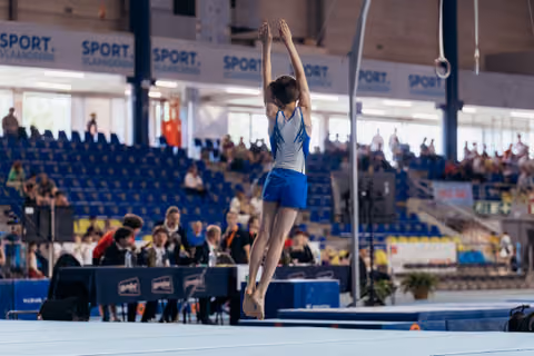 Young gymnast mid-air with arms raised overhead, landing on floor exercise mat in a packed indoor arena.