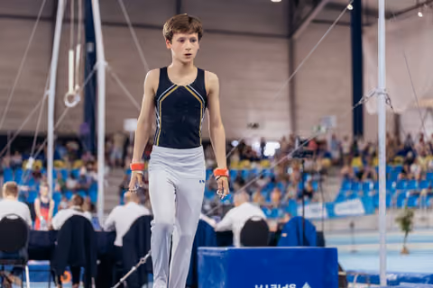 Young male gymnast walks with focused expression across the floor, wearing black leotard and white pants in a busy sports arena.