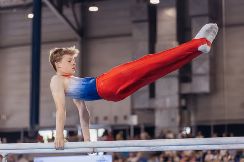 Young male gymnast performs a straddle hold on parallel bars, expression focused and determined, indoor arena.