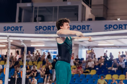 Young male gymnast grips parallel bars with intense grimace, wearing green and black leotard, crowd watching in background.
