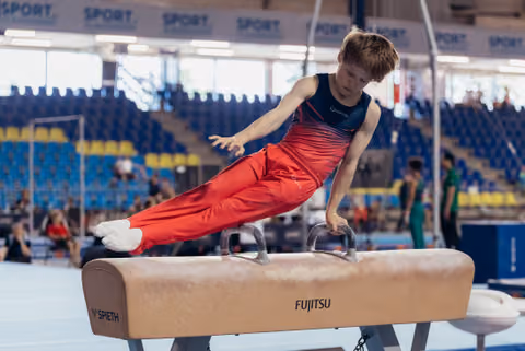 Young male gymnast performs a focused pommel horse routine in a red and navy leotard at an indoor sports arena.