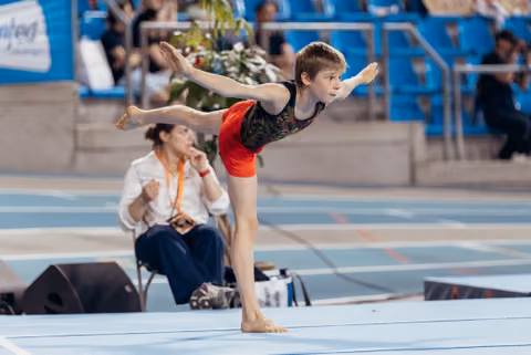Young male gymnast performs a focused arabesque balance on floor, arms extended, judge seated behind him.