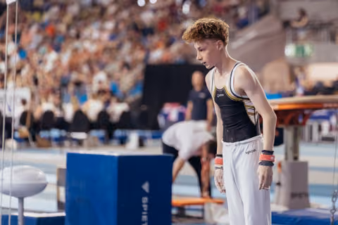 Young male gymnast with red hair leans forward, focused and composed, studying the vault before his attempt in a packed arena.