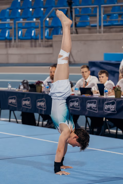 Young gymnast performing a straight handstand on floor, judges seated at Gymfed table in background.