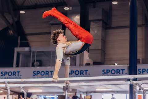 Male gymnast performs an inverted pike hold on parallel bars, legs raised in red uniform, intense concentration indoors.