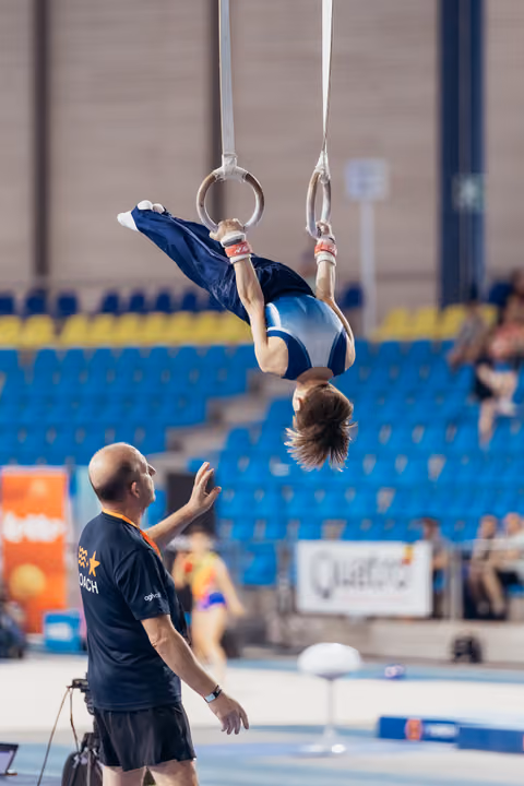 Young gymnast hangs inverted on still rings while attentive coach below reaches up to guide and spot during an indoor gymnastics event.