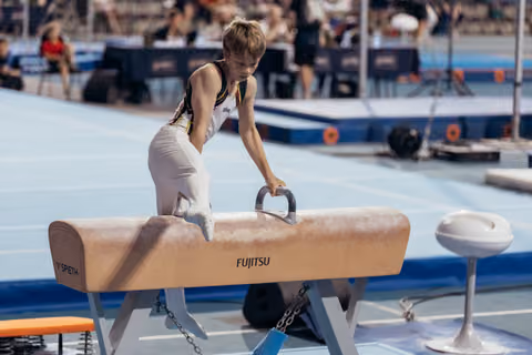 Young male gymnast leans intently over a Fujitsu pommel horse, gripping the handles with focused concentration at an indoor sports event.