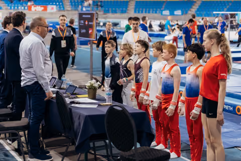 Young male gymnasts in leotards line up attentively before officials at a table during an indoor gymnastics competition.