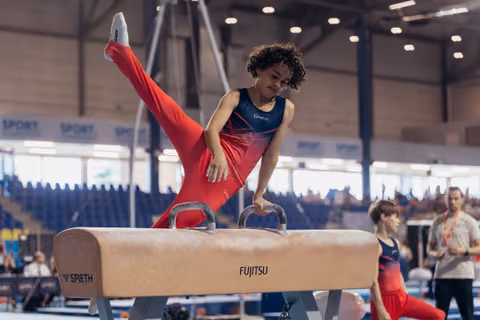 Young gymnast in red performs a focused routine on the pommel horse, one leg raised high in a competition hall.