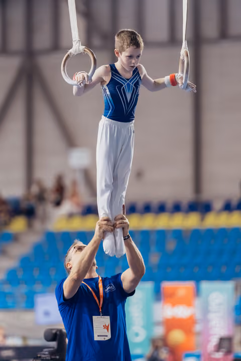 Focused young gymnast holds still rings while coach steadies his feet in a blue-seated sports arena.