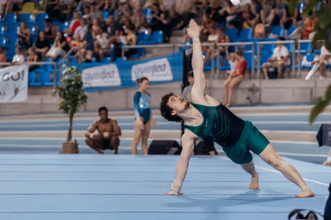 Male gymnast in teal leotard performs a dramatic side stretch on floor, arm raised high, focused gaze upward in a packed arena.
