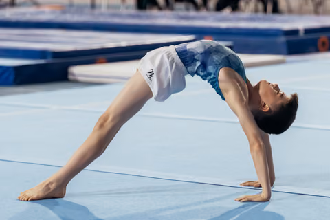 Young gymnast performs a back bridge on a blue gymnastics mat, arching gracefully with a focused expression.